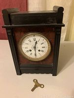 Front view of the antique tabletop grandfather clock with white Roman numeral dial, brass bezel, and black wooden case with carved sides. Brass winding key placed in front.