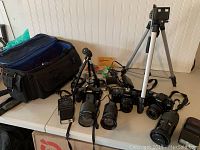 Wide photo displaying three Minolta SLR cameras, assorted lenses, four flashes, two tripods, and a large camera bag with accessories on a white table.