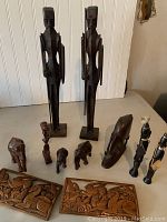 Front view of wood carved items showing two tall human figures, smaller animal and human figurines, and carved wooden panels