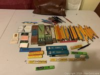 Top-down photo showing the full collection arranged on a white cloth surface, including the bag, pencils, drawing leads in boxes, erasers, injectors, and stamp pad.