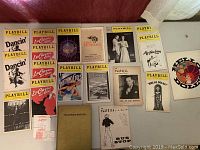 Photo showing a collection of vintage Playbills and programs laid out on a surface, including titles like Dancin', La Cage aux Folles, Anything Goes, and additional memorabilia such as the Hollywood Babylon first edition book.