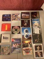 Photo showing 18 rock CDs and DVDs laid flat on a surface, including Pink Floyd Pulse DVD, multiple Beatles CD compilations, Aerosmith and Ozzy Osbourne CDs.
