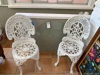 Two white painted cast iron garden chairs with ornate scroll and floral lattice designs and a round glass tabletop leaning against the wall.