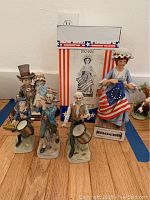 Porcelain figurines grouped with Betsy Ross holding flag, colonial drummers, and large Betsy Ross box in background
