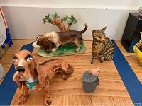 Four ceramic animal figurines on wooden surface with two Basset Hound dog figurines, one tabby cat, and one small cat on a trash can.