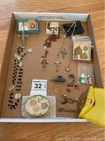Top-down photo showing multiple rosaries, crosses, small pins, medals, and a vintage face powder box on a cardboard surface.