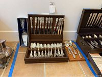 Brown storage box holding 12 dinner knives standing upright in the top compartment, 12 dinner forks and 12 teaspoons lying flat in the bottom compartment, all with ornate silver plated handles. Five additional serving pieces (forks and spoons) lie beside the box on the floor.