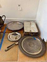 Full view of the chafing dishes, serving platters, butter knife and ice cream scoop arranged on the floor. Tarnishing visible, handle broken on one chafing dish.