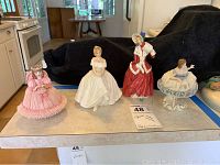 Four porcelain women figurines displayed on a surface under natural light, showing variety in dresses and postures