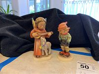 Two Hummel porcelain figurines on a table with a black cloth background. Angel figure with lamb and boy figurine standing beside each other.