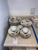 Photo showing stacks of Royal Worcester plates, mugs, and a bowl with fruit designs and gold trim on a countertop.