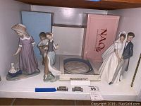 Four Lladro porcelain figures and a decorative plate displayed on a white shelf with boxed packaging partly visible behind items.