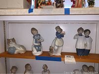Front view of all four Lladro figures on wooden shelf showing the cat, girl with teddy bear, woman with basket and small dog, and two children figures.