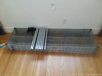 Stack of flat wire metal shelves with supporting metal poles and black plastic foot caps on hardwood floor.