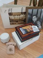 Photo showing two vintage framed photos, ceramic rotary telephone music box, wooden music box and white box on a wooden surface