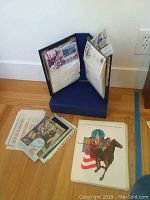 Photo of multiple stamp albums and folders on a wooden floor, showing a partially open album with protective sleeves containing stamps and covers, and loose folders and sheets nearby.