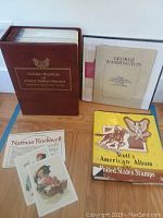 Large brown binder titled 'Golden Replicas of United States Stamps' alongside Norman Rockwell lithographs and Scott's American Album book.