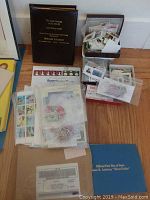 Photo showing a collection of assorted postage stamps in sleeves and envelopes, a gold stamp album, and other related materials on the floor.
