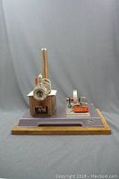 Front and side view of the vintage Wilesco miniature steam engine showing the boiler with brick pattern, chimney, and engine flywheel on a purple-grey base with wooden edges.