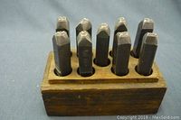 Set of 11 vintage metal number stamps inserted vertically into a wooden block holder against a blue fabric background. Stamps appear dark metal with pointed ends and squared tops.
