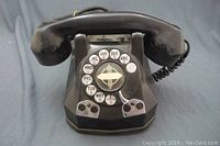 Front view of vintage black rotary phone showing handset, rotary dial with numbers and letters, and coiled cord.