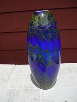 Front view of a tall cobalt blue glass vase with yellow and green wave designs against a red wooden background on a white surface.