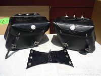 Pair of black leather motorcycle saddlebags shown from front with silver conchos, two front buckle straps, and a detached black leather center connector piece with snaps in front of the bags.
