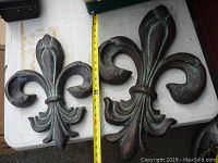 Two fleur-de-lis wall mounts side by side on a white table with measuring tape showing length
