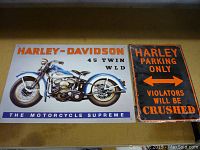 Front view of two Harley Davidson metal plaques on a surface. The first plaque features a painted vintage motorcycle image and text, the second plaque a large text warning.