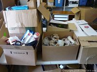 Wide view of three large cardboard boxes filled with assorted decorative and houseware items including ceramic figures and household objects, boxes placed on a table.