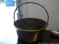 Full view of the antique heavy brass pot showing the entire exterior and attached handle, placed on a flat surface in a workshop or garage.