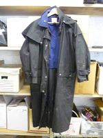 Full front view of the black heavy duty raincoat hanging on a hanger. Visible details include the cape-style shoulder flap, button closures, and long silhouette.