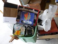 Top view of large green bin filled with boxes and miscellaneous items including a book about paper airplanes and plastic bags