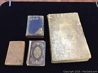 Four antique books displayed on a black cloth background showing wear and damage on covers and spines.