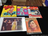 Overview photo showing all five ephemera items displayed on a floor wooden surface: Bette Midler tour booklet, Elvis paper doll book, Shredded Wheat Wayne Gretzky box, All About Eve lobby card, and Screen Book magazine 1935.