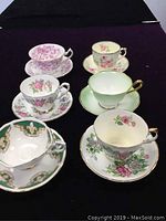 View of all six vintage bone china cups and saucers arranged on black background, showing floral patterns and gold trims.