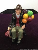 Front view of the vintage ceramic figurine of a seated man holding colorful balloons and a sack.