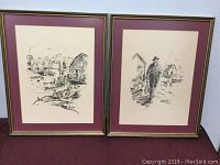 Both charcoal drawings framed and displayed side by side against a white wall and burgundy surface.