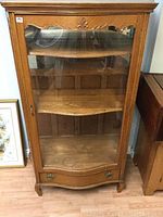 Front view of vintage oak display cabinet showing bow-front glass door, one drawer with brass handles, and wood carvings on top edge.