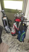 Wide shot of three golf bags each containing golf clubs, displayed side-by-side adjacent to a window on a concrete floor.