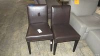 Two brown leather chairs placed side-by-side on concrete floor, showing front and side views.