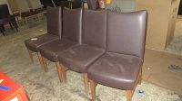 Four matching brown leather chairs with wooden legs arranged side by side on concrete floor, showing overall condition and wear.