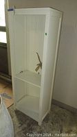 Photo of tall cream colored bookshelf showing front and side with three shelves, set against wall.