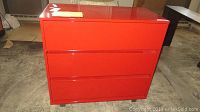 Front view of a red lacquered dresser with three drawers, showing a smooth glossy finish and integrated drawer handles.