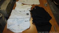 Overview of 4 white laundered non-matching chef jackets and 7 black embroidered matching chef jackets spread on wood table.