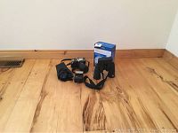 Wide shot showing all items in the lot including binoculars, camera body, lens, flash, and wifi extender box on wooden floor.