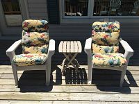 Two white plastic Muskoka chairs with floral cushions and a small white folding side table arranged on a wooden deck.