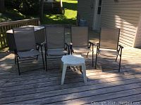 Four grey mesh and metal folding chairs positioned in a row with a white plastic side table on a wooden deck, showing outdoor setting.