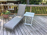 Outdoor reclining lounge chair with accompanying small folding side table positioned on a wooden deck with wooden railing and potted plants visible.