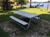 Full view of white plastic top picnic table with attached benches and metal frame set outdoors on gravel near water.